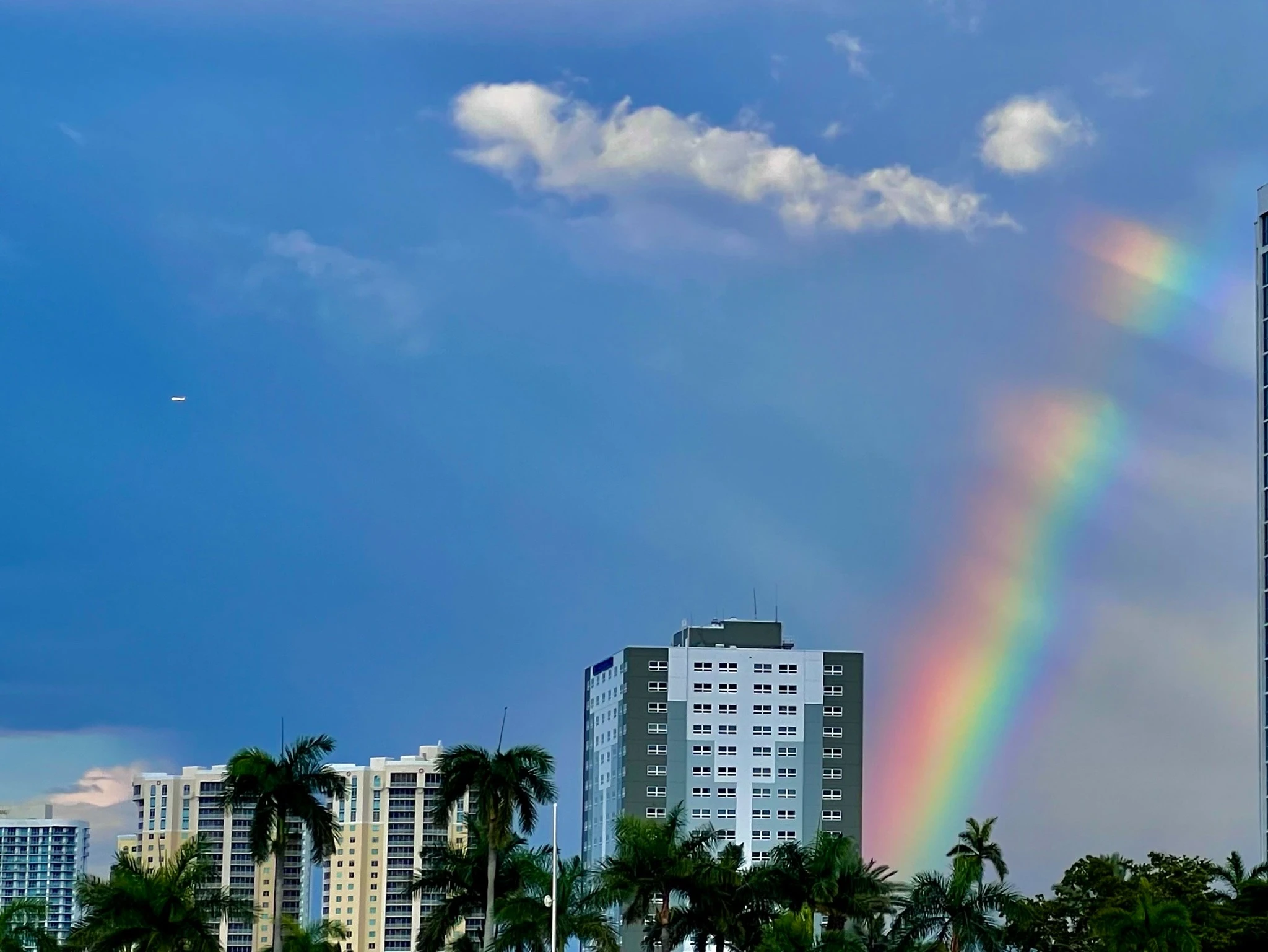 A rainbow appearing in separated sections through passing clouds above city buildings and palm trees